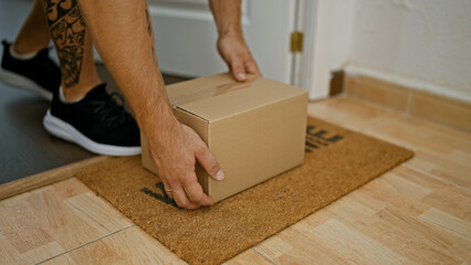 A tattooed man picks up a cardboard box from a doormat inside a home, signifying delivery or moving.