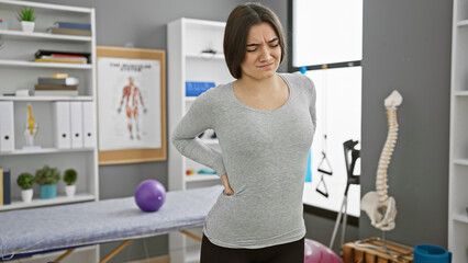 A young hispanic woman experiencing back pain stands in a physiotherapy clinic with anatomy posters...