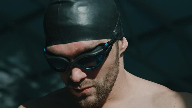 Close-up portrait shot of calm, confident young Caucasian male athlete with stubble and beard, in black rubber cap putting on goggles and looking away, while getting ready for competition in pool