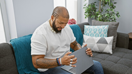 African american man closing laptop at home, with a beard and tattoos, showcased in a modern interior setting.