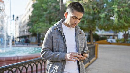Obraz premium A young hispanic man in casual attire is focused on his smartphone while standing outdoors in a city park.