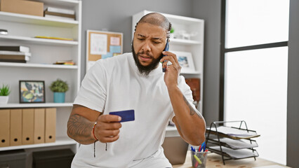 A concerned adult black man holds a credit card and talks on the phone in a modern office setting.