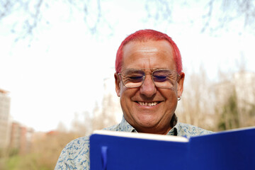A senior man with painted hair and glasses is smiling while reading a book in a park