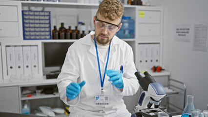 A young bearded caucasian man in a laboratory, wearing safety goggles and blue gloves, analyzing a...