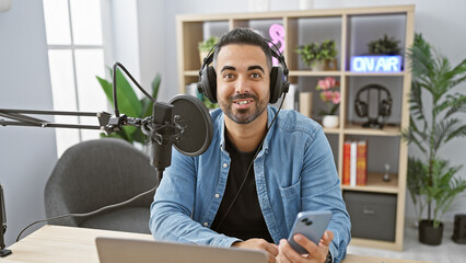 Handsome hispanic man with a beard smiling while hosting a show in a radio studio interior.