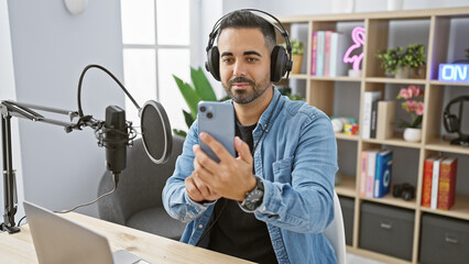 Bearded hispanic man in studio with microphone and headphones holding smartphone, embodying...