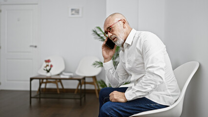 Bald man with beard and glasses, using a smartphone, sits in a modern white room with a wooden...