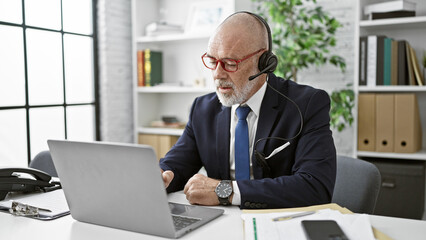 Fototapeta na wymiar A professional senior man with beard and glasses works attentively in a modern office using laptop and headset.