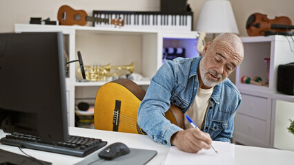 A mature man writing in a creative indoor workspace with musical instruments and computer equipment.
