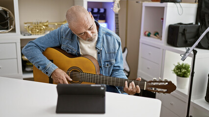A mature man plays guitar while watching a tutorial on a tablet in a well-lit home office.