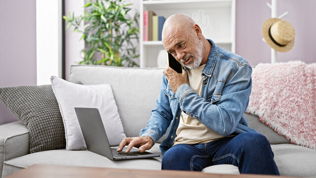 Bald man with beard using laptop and talking on phone in modern living room. - Powered by Adobe