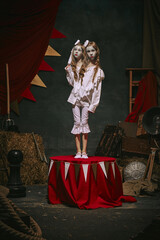 Siamese twin girls in white vintage costumes with makeup stand on stage on dark retro circus...