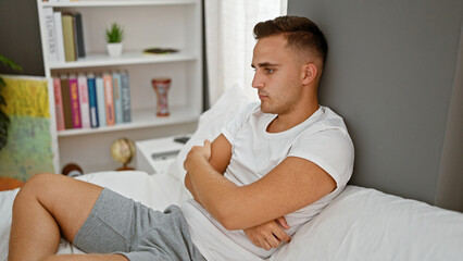 A young man in casual attire sits thoughtfully in a modern bedroom with bookshelf and decorative...