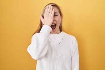 Young caucasian woman wearing white sweater over yellow background covering one eye with hand, confident smile on face and surprise emotion.