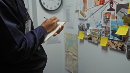 An african american man takes notes at a police station with an evidence board in the background,...