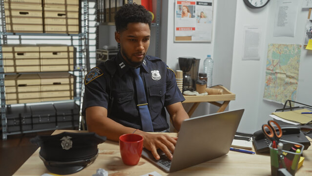 An african american male police officer analyzes evidence on a laptop in a crowded detective's office.