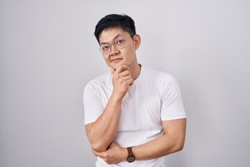 Young asian man standing over white background with hand on chin thinking about question, pensive expression. smiling with thoughtful face. doubt concept.