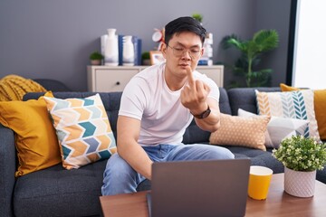 Young asian man using laptop at home sitting on the sofa showing middle finger, impolite and rude...
