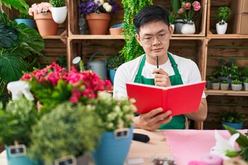 Young chinese man florist reading notebook with doubt expression at flower shop