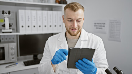 Naklejka na ściany i meble A young caucasian man in a white lab coat and blue gloves examines a tablet in a modern laboratory setting.