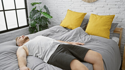 Young caucasian man relaxing on bed in a modern bedroom with white walls, yellow pillows, and green...