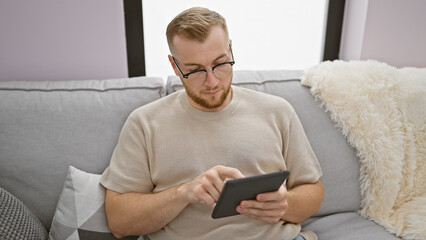 A young bearded man in glasses intently uses a tablet on a cozy sofa in a modern living room.