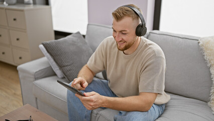 Bearded young man with headphones using tablet while sitting on couch indoors.