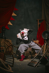 Clown in black hat, white face with red nose and striped pants shouting over dark retro circus...