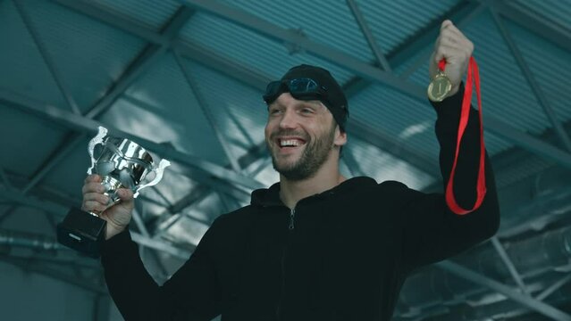 Medium close-up shot of cheeful Caucasian male athlete in cap, goggles, zip hoodie, clutching gold medal and kissing trophy cup while posing for photos in pool after winning swimming competition