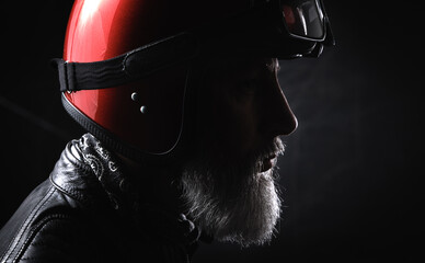 Portrait d'un motard homme avec un casque rouge et une veste en cuir et des lunettes de protection et une barbe grise vintage - 764952498