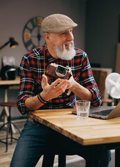 Portrait d'un homme assis photographe hipster situé dans un studio atelier et qui tient un appareil photo vintage en souriant devant un ordinateur - 764952633