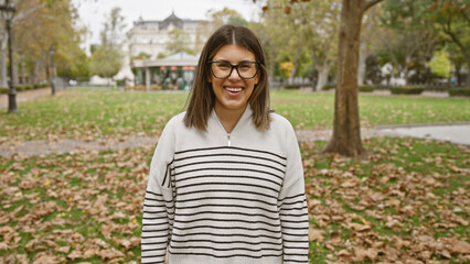 A smiling young hispanic woman stands in an autumnal park wearing glasses and a striped sweater.