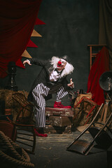 Clown in black hat, white face with red nose and striped pants playing over dark retro circus backstage background. Comedian. Concept of circus, theater, performance, show, retro and vintage