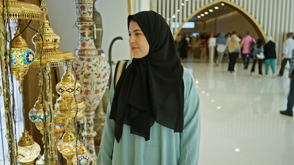 A smiling woman in a hijab admires ornate lanterns at a bustling abu dhabi shopping mall.