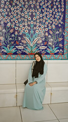 A smiling young woman in a hijab sitting in front of an ornate floral islamic background at a mosque in the emirates.
