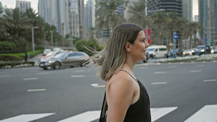 Obraz na płótnie Canvas A young brunette woman wearing casual clothing stands by a crosswalk amidst the modern skyscrapers of dubai's cityscape.