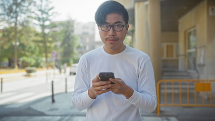 A young asian man uses a smartphone while standing on a city street, conveying urban connectivity.