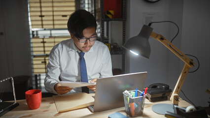 Focused asian man investigating crime evidence in a dimly lit detective office.