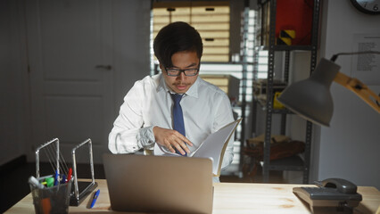 Asian man in a detective office analyzing documents at night, illuminated by a desk lamp.