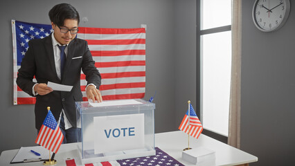 Young asian man in a suit voting indoors with us flags and ballot box, displaying civic duty and...