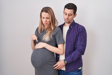 Young couple expecting a baby standing over white background pointing down looking sad and upset, indicating direction with fingers, unhappy and depressed.