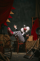 Clown in black hat, white face with red nose and striped pants performing over dark retro circus...