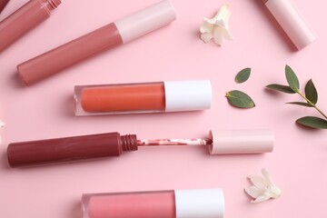 Different lip glosses, applicator, flowers and green leaves on pink background, flat lay