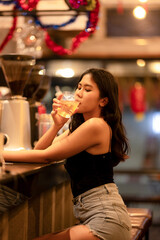 A young woman is sipping a cold alcoholic drink. While sitting at the bar in a rustic cafe and looking at camera.