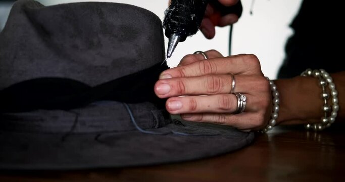 A close-up of the process of decorating a fancy hat. A woman hatmaker uses a glue gun to decorate a hat with feathers, threads, and fabric. Fashion designer workplace.