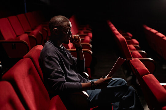 Side view portrait of African American man sitting in audience and reading script in empty theater copy space