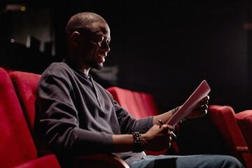 Side view portrait of smiling Black man sitting in audience and reading script in low light at...