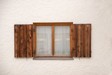 closeup of a window with brown wooden shutters and white curtain, plastered concrete facade