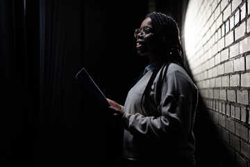 Minimal side view portrait of Black young woman rehearsing on stage in theater with low light copy...