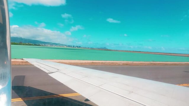 A plane is moving along the runway in Oahu airport, ready to take off. View through the airplane window as the plane is speeding up to take off. Departure of a plane from a tropical island with ocean,
